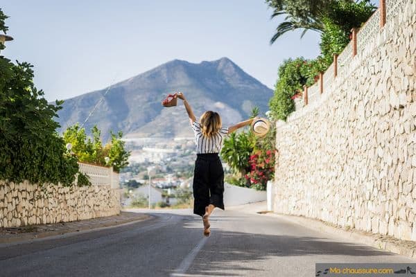 mujer caminando por la calle sosteniendo sus zapatos de verano en sus manos